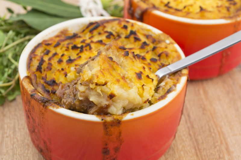 Individual cottage pies topped with white and sweet potato mash on a wooden surface.